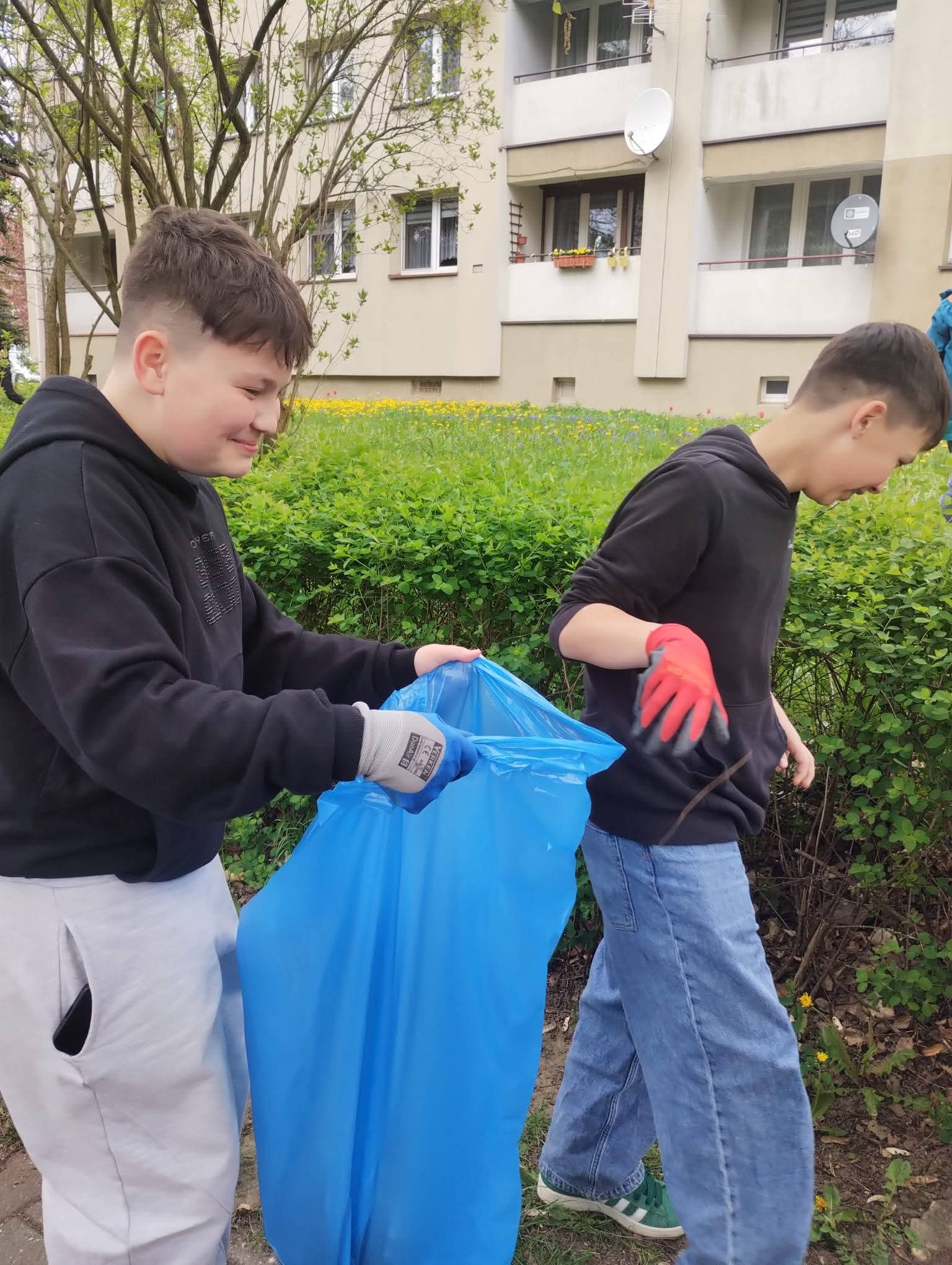 Dwóch chłopców zbiera śmieci do niebieskiego worka podczas sprzątania terenu zielonego.