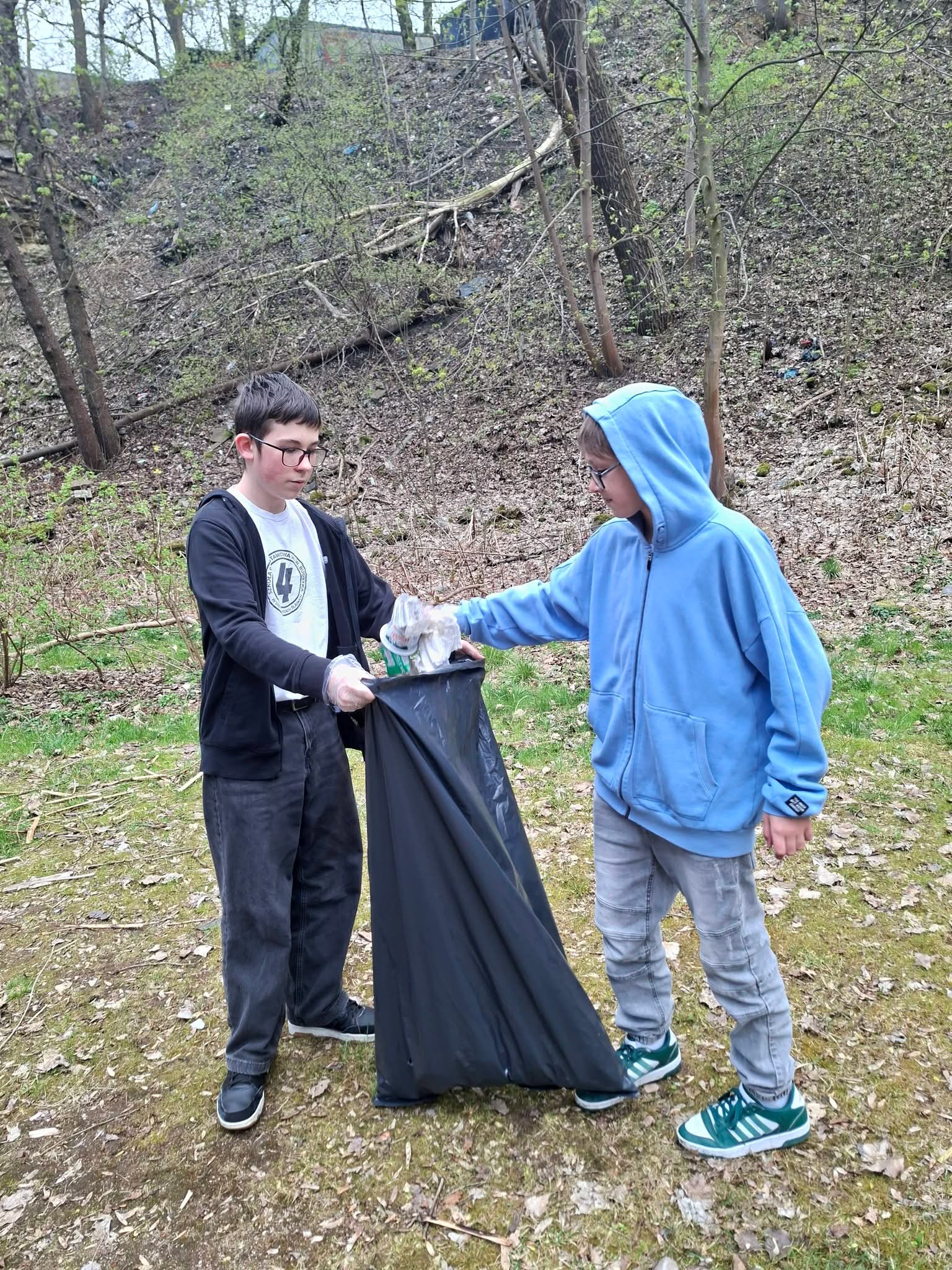 Dwóch chłopców zbiera śmieci do czarnego worka w lesie.