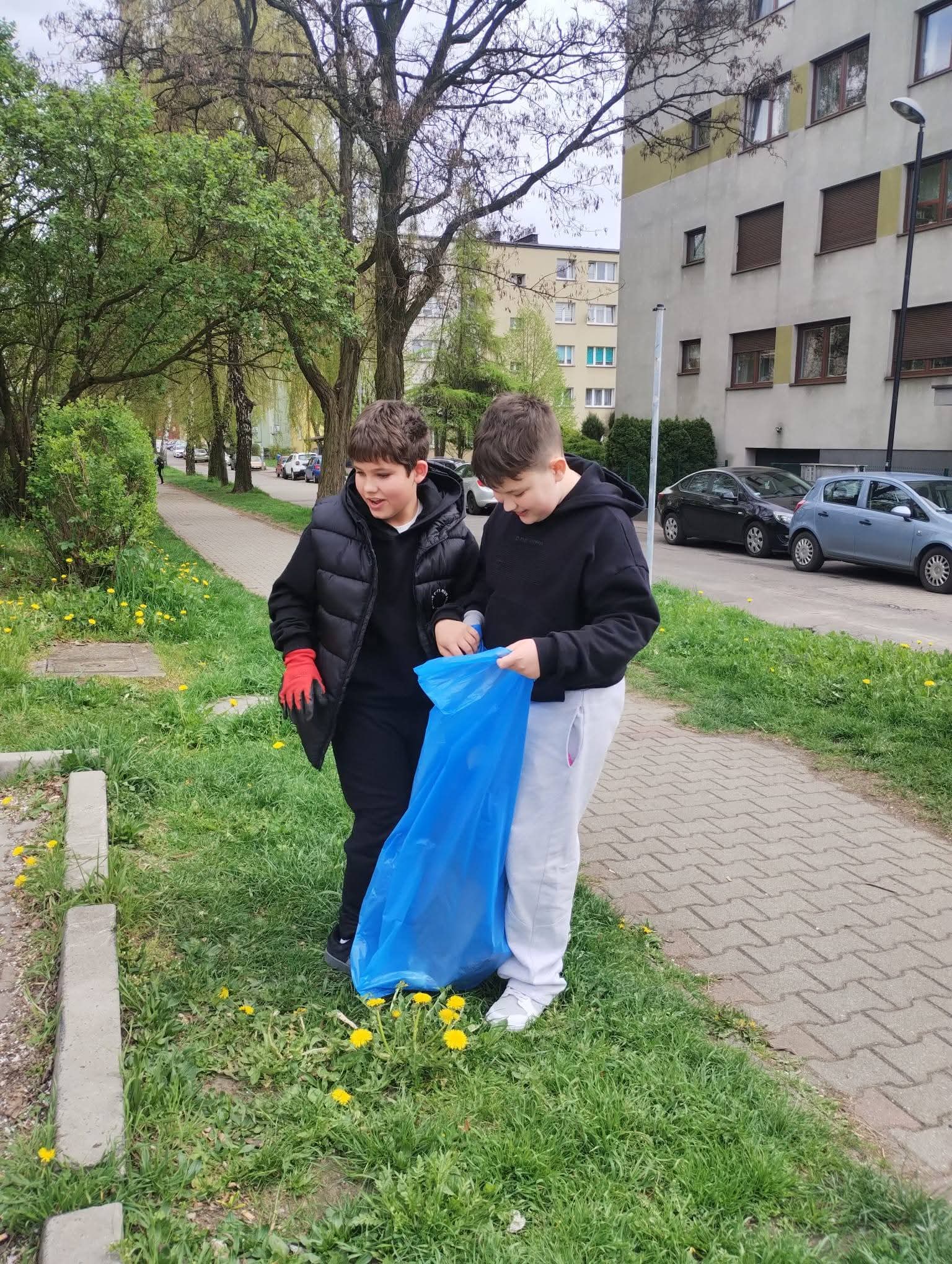 Dwóch chłopców zbiera śmieci do niebieskiej torby na trawniku z żółtymi kwiatami.