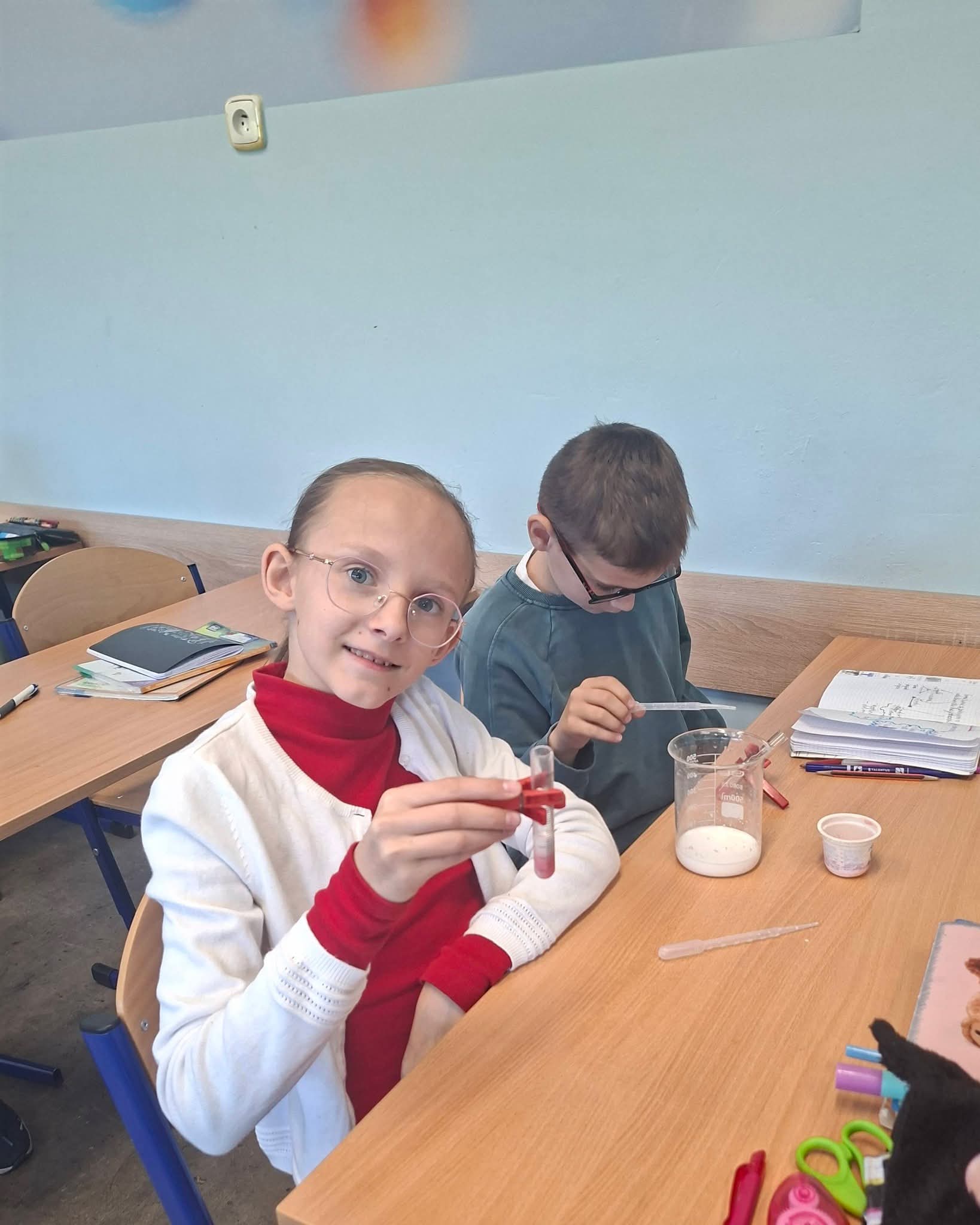 Dzieci prowadzą eksperymenty chemiczne w klasie, trzymając probówki i pipety.