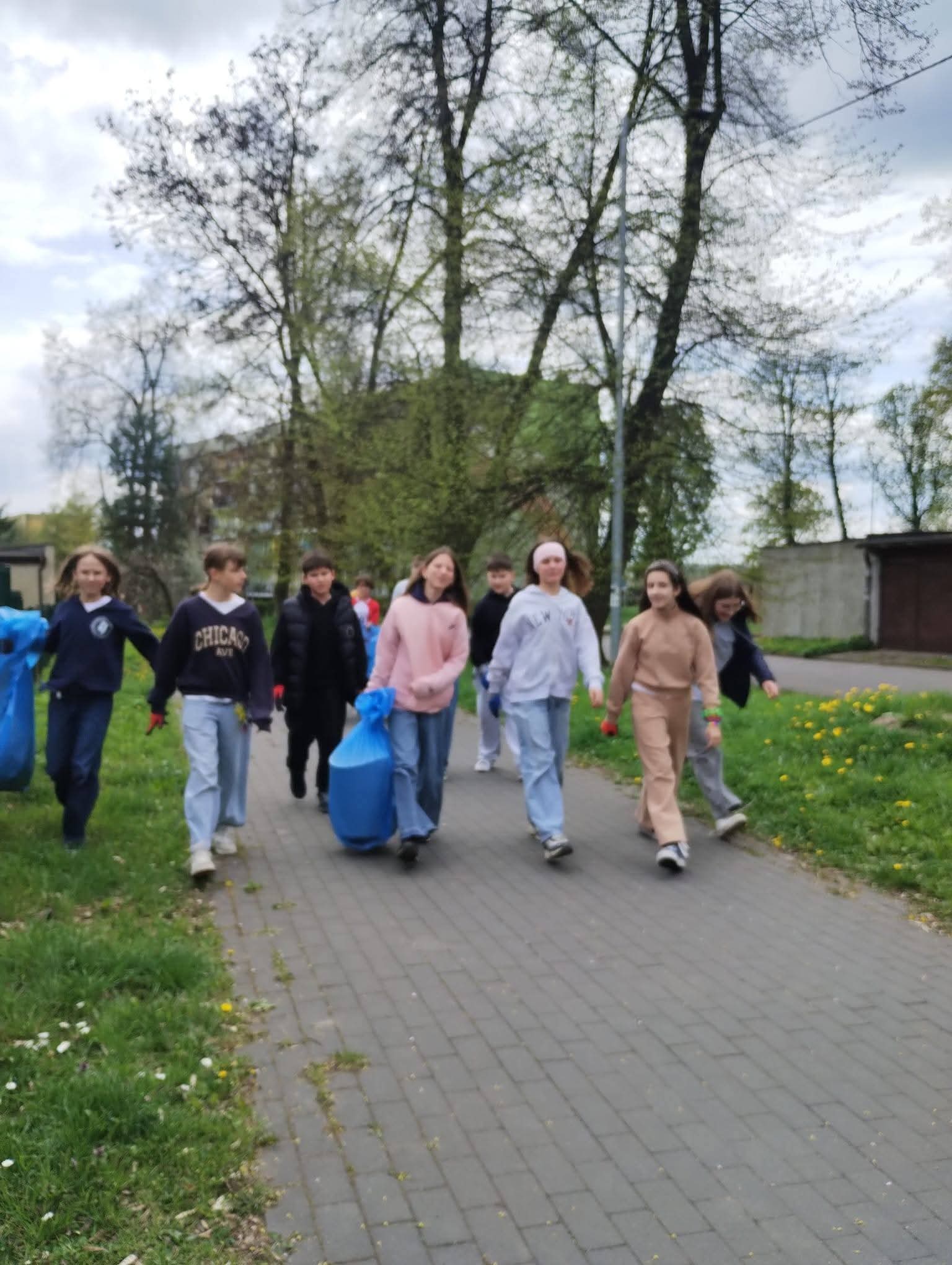 Grupa dzieci idzie chodnikiem z workami na śmieci, sprzątając teren.