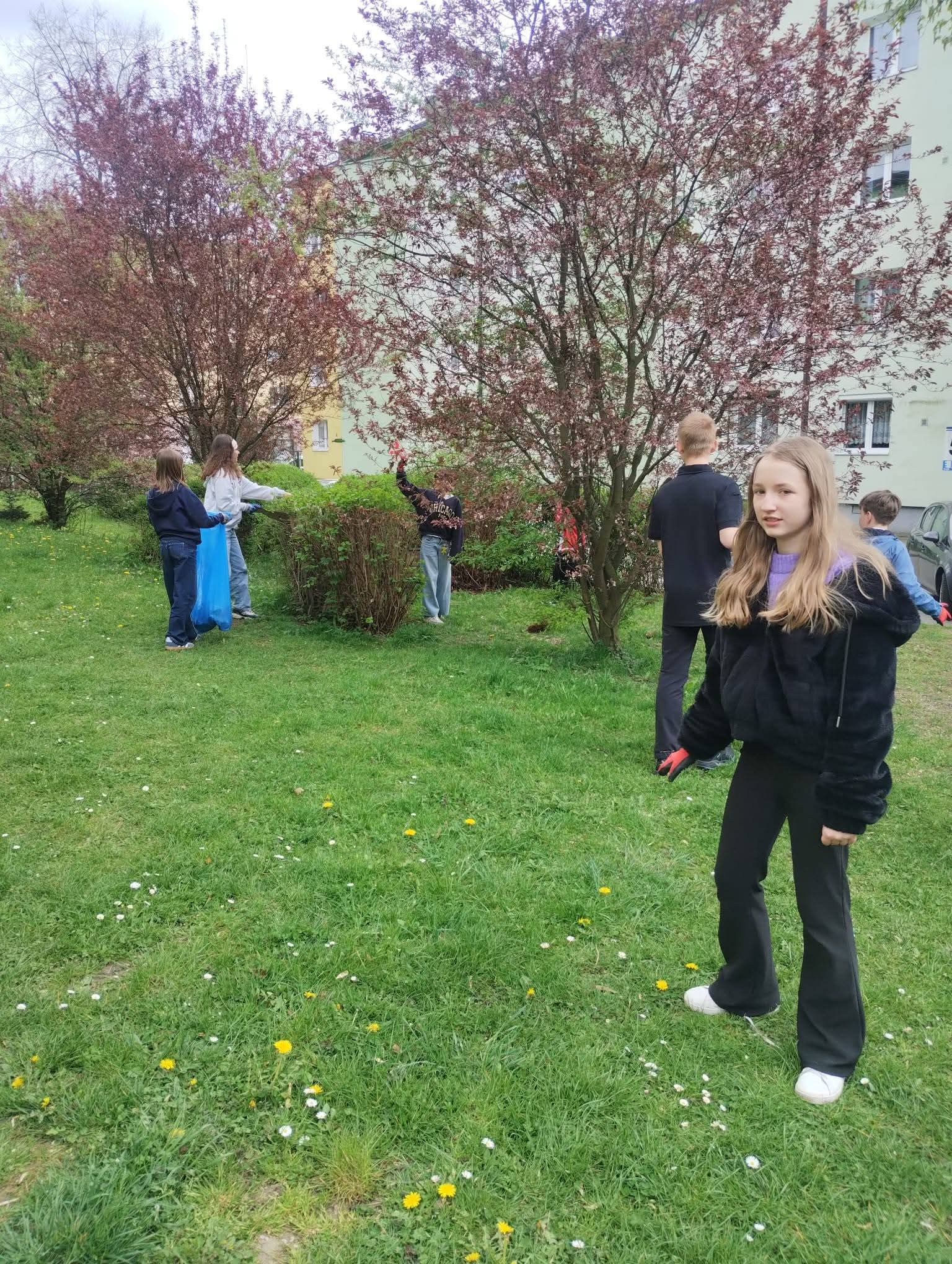 Grupa dzieci bawi się na trawiastym podwórku z drzewami w tle.