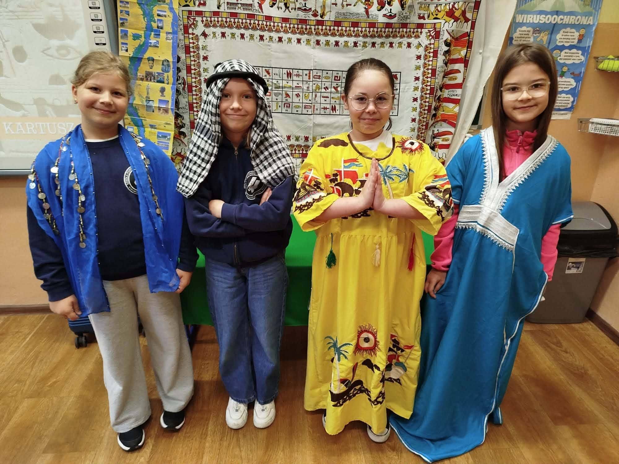 Cztery dziewczynki w strojach ludowych z różnych kultur stoją przed mapą świata.