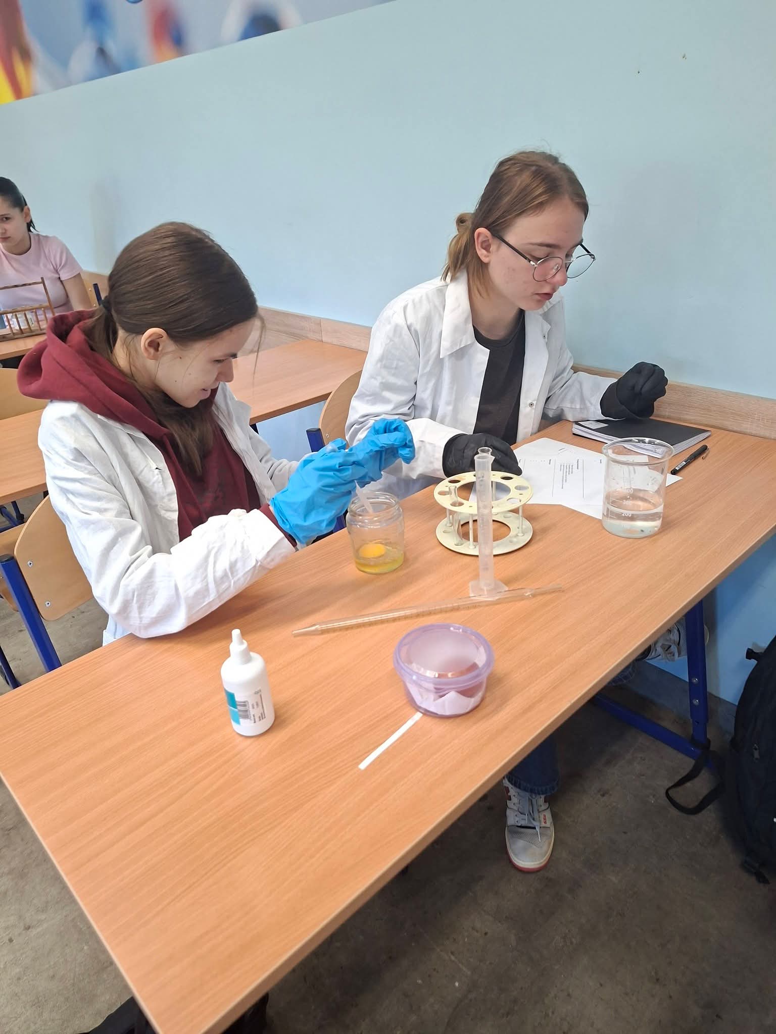 Dziewczyny w fartuchach laboratoryjnych wykonują eksperymenty chemiczne przy biurku.