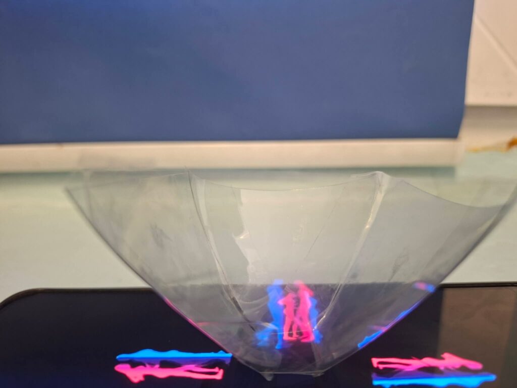 Hologramowa piramida na smartfonie wyświetla trójwymiarowe postacie w kolorach niebieskim i różowym.