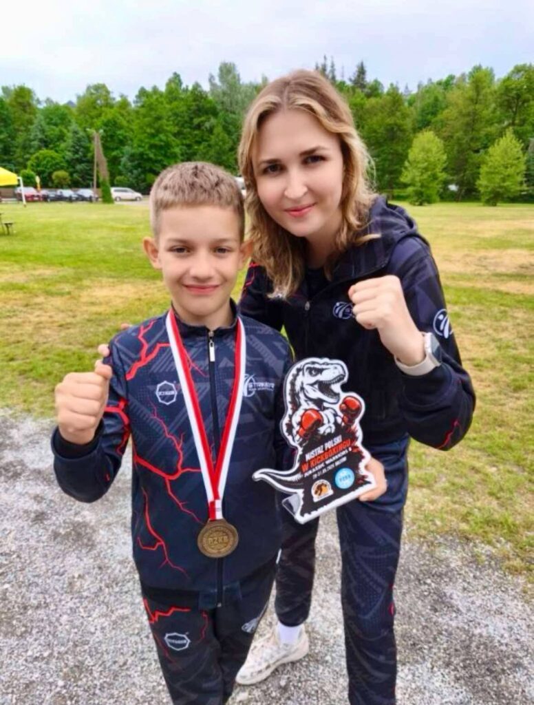 Uśmiechnięty chłopiec w sportowej bluzie, z medalem na szyi, trzyma statuetkę z dinozaurem i napisem „Mistrz Polski w Kickboxingu”. Obok niego stoi dumna kobieta (trenerka lub mama). Oboje pozują z uniesionymi w geście walki pięściami na tle zielonego parku.