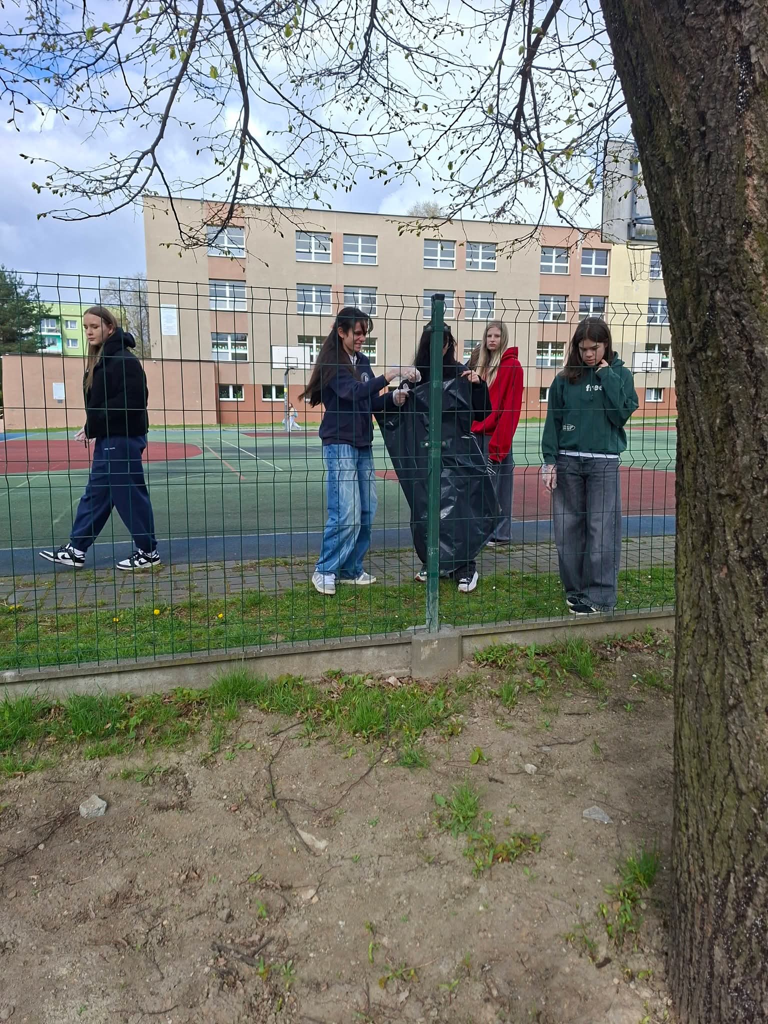 Młodzież sprząta teren wokół boiska szkolnego, zbierając śmieci do czarnego worka.