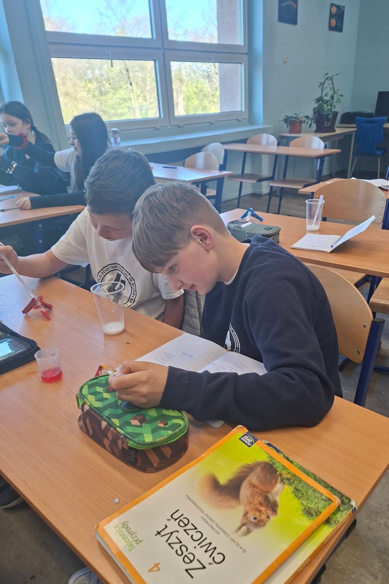 Chłopcy prowadzą doświadczenie chemiczne w klasie, patrząc na zeszyty i płyny.