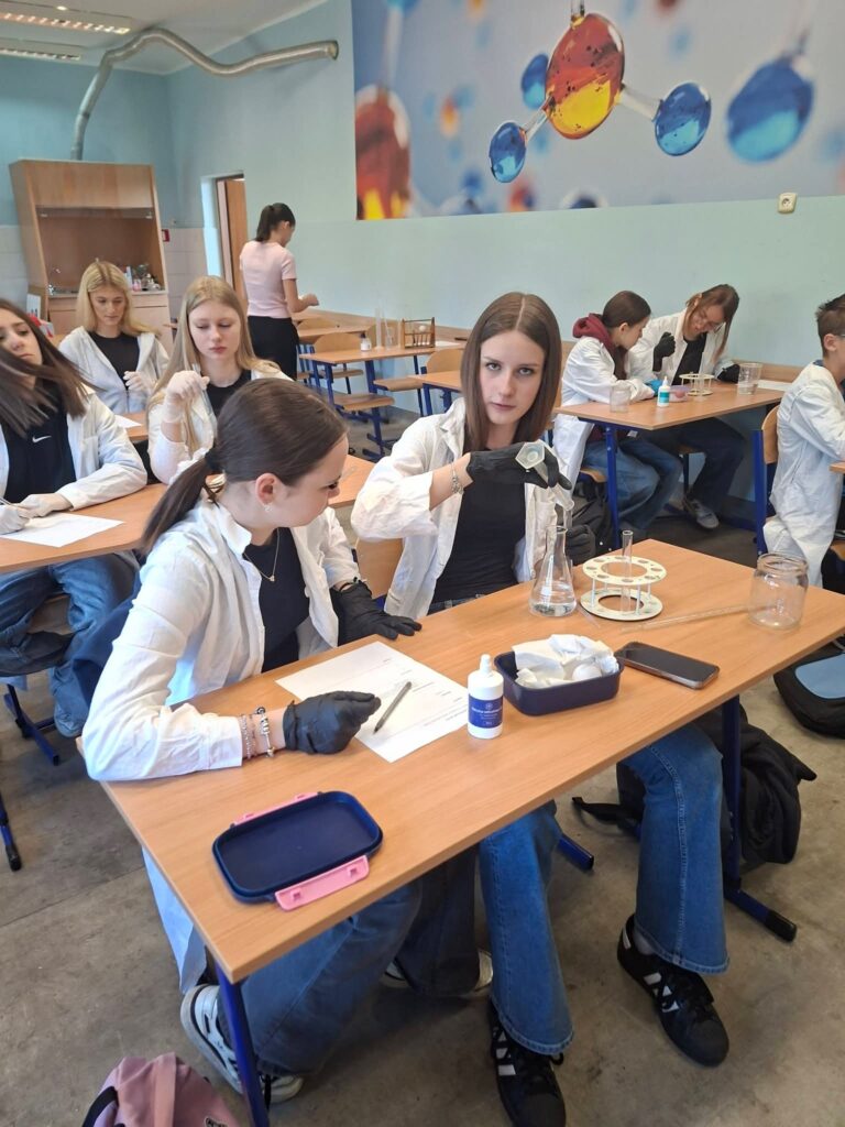 Uczennice w fartuchach laboratoryjnych przeprowadzają eksperymenty chemiczne przy biurkach.