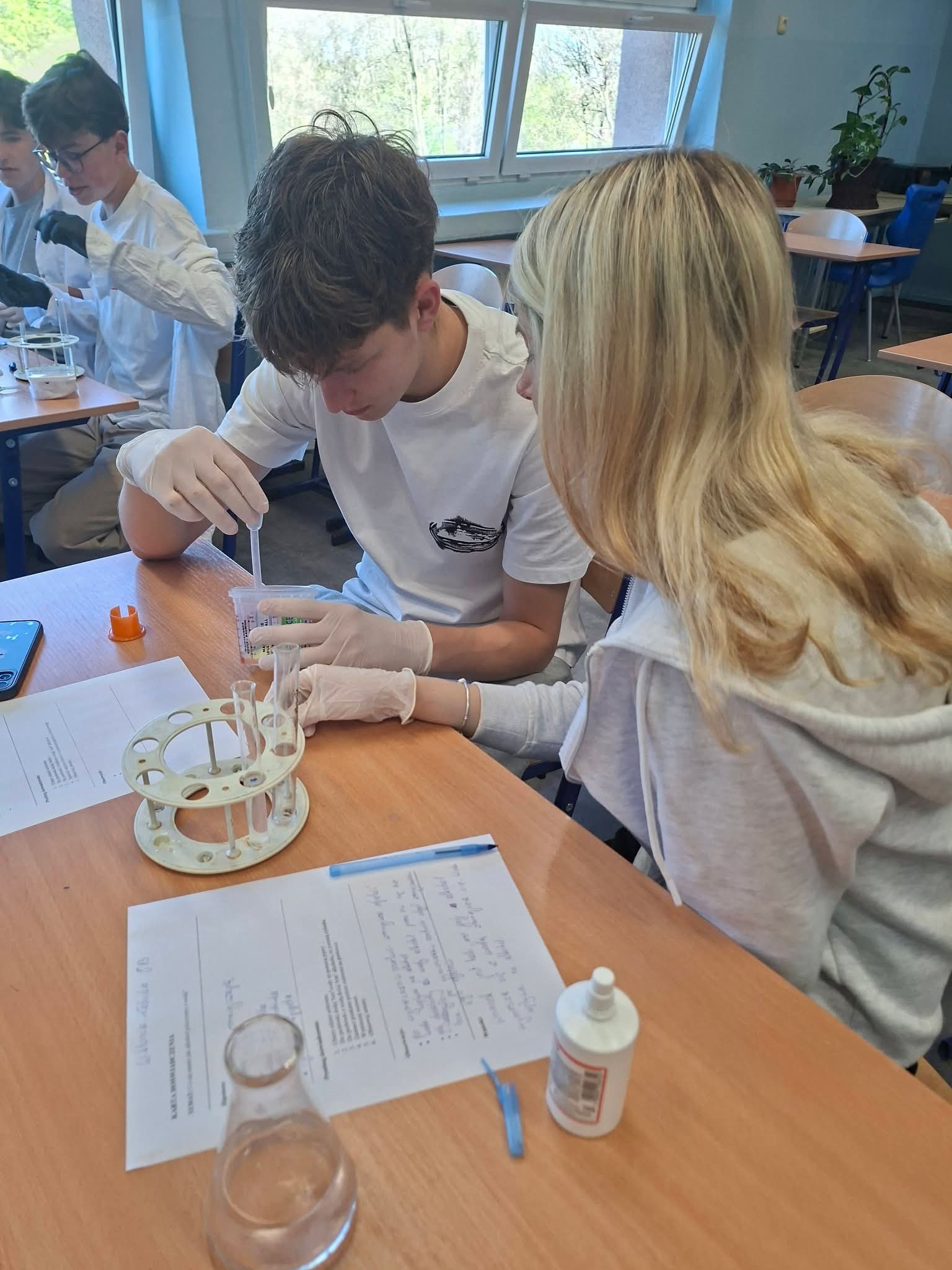 Uczniowie w fartuchach laboratoryjnych wykonują eksperyment chemiczny w klasie.