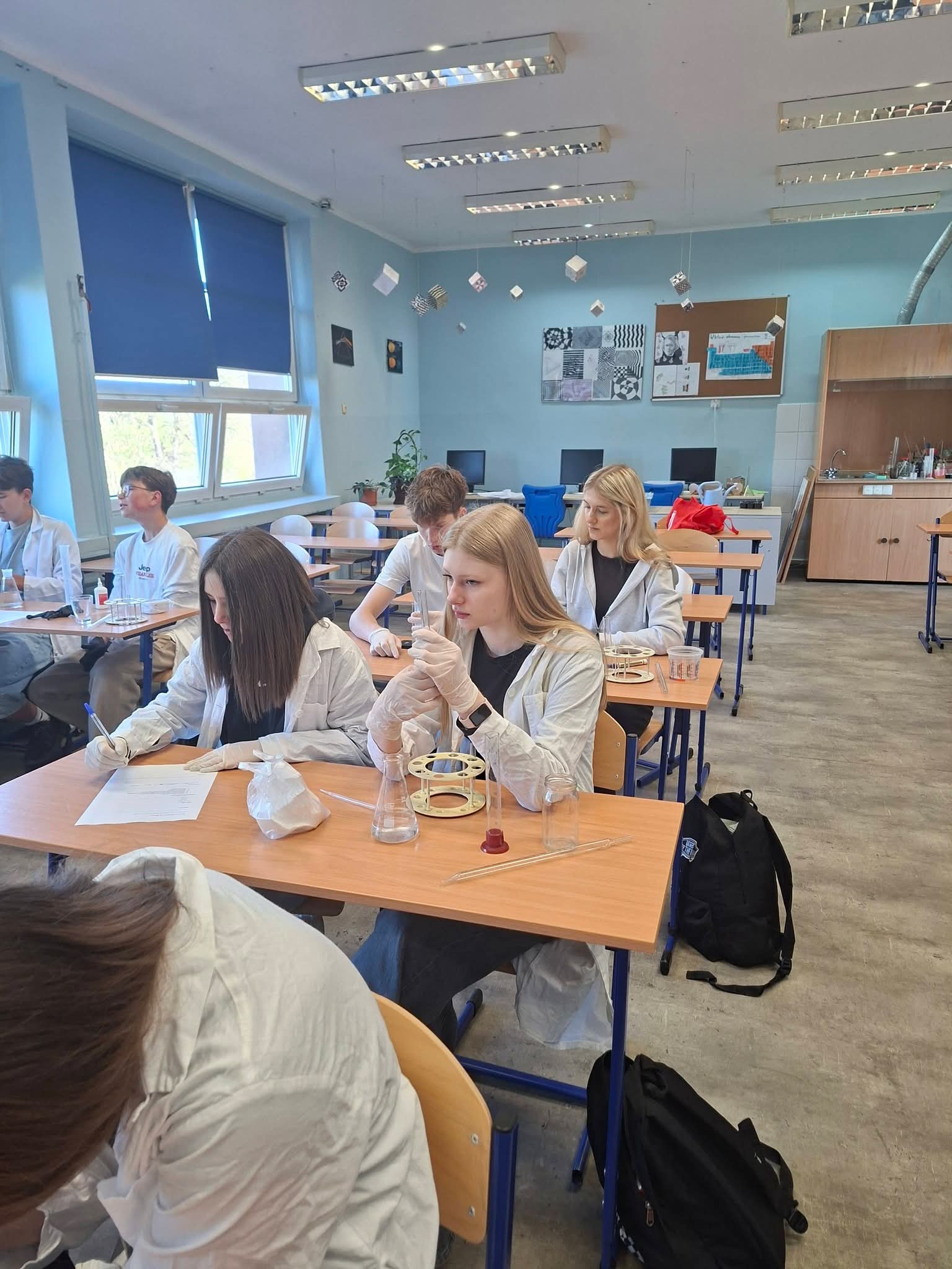Uczniowie w fartuchach laboratoryjnych wykonują eksperymenty chemiczne przy biurkach.